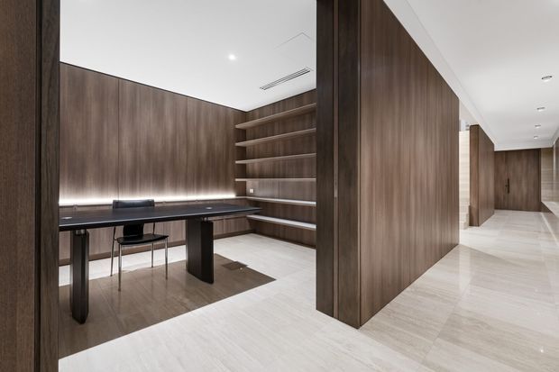 A refined contemporary interior featuring a compact study nook or home office enclosed by dark timber wall panelling. The space includes a black desk, simple chair, integrated open shelving with concealed LED lighting, and pale stone-look flooring. A bright hallway with matching timber panels and recessed ceiling lights extends to the right.