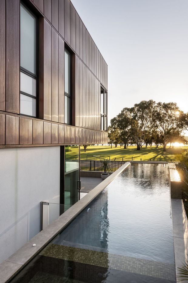 Contemporary two-storey building with a projecting upper volume clad in dark timber-toned vertical panels and tall black-framed glazing. A long narrow reflecting or swimming pool runs alongside the building, overlooking a manicured open lawn and mature trees in warm late-afternoon light.