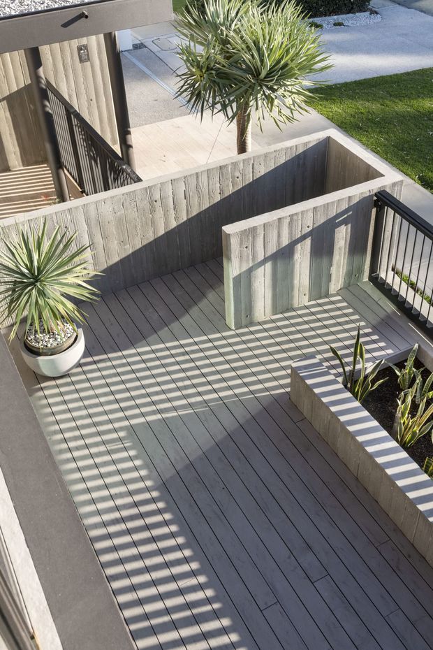 Elevated view of a contemporary outdoor terrace or deck with dark timber decking, grey weathered timber privacy screens and raised planter beds. Black metal railings, potted spiky plants and a paved garden path surround the deck, with strong sunlight casting linear shadows across the space.