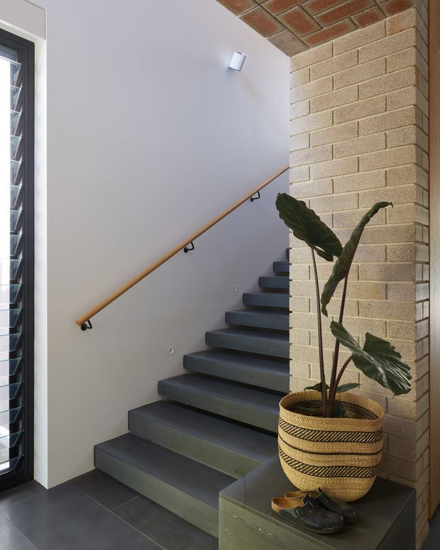 Interior stairway with dark tiled or concrete treads, a simple timber handrail, white plaster walls, and exposed light-coloured brickwork forming a feature wall and ceiling soffit. A tall louvre window brings in daylight, while a woven planter and greenery add warmth beside the stairs.