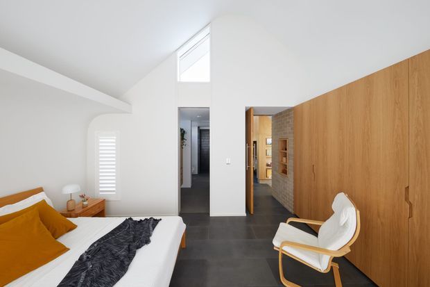 Bright contemporary bedroom with a low-profile bed dressed in white and mustard bedding, a dark textured throw, a timber bedside table, and a sculptural white lounge chair. A full-height timber built-in storage wall lines the right side, while dark polished flooring, white walls, a high clerestory window, and an open doorway create a clean, airy interior.