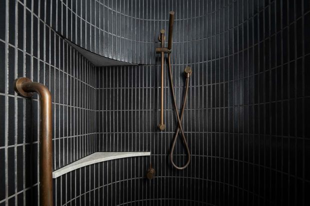 A compact, dark shower enclosure lined with glossy black rectangular tiles and contrasting pale grout. Exposed copper-toned plumbing and a wall-mounted handheld shower set create an industrial, tactile character. A small angular tiled ledge runs along the left wall.