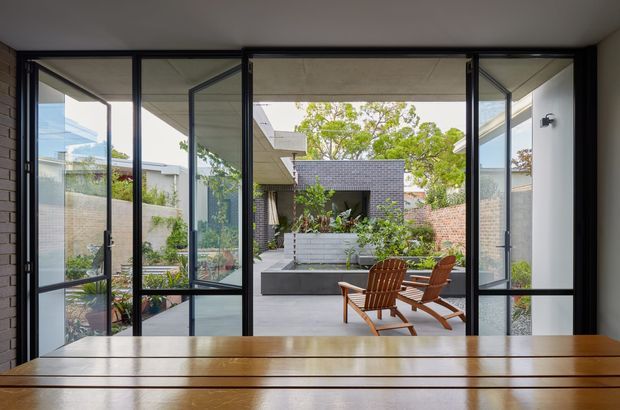 View from an interior dining area through expansive black-framed, bi-fold or folding glass doors into a landscaped courtyard. A timber dining table dominates the foreground, while timber outdoor lounge chairs, raised planters, rendered and brick walls, and a partially covered courtyard are visible beyond. The composition highlights indoor-outdoor connection and generous glazing.
