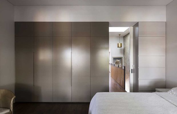A refined contemporary bedroom with a large wall of dark built-in wardrobe cabinetry, a partially visible bed with white textured bedding, dark timber flooring, and a concealed doorway leading toward a warmly detailed adjacent space. The palette is subdued and neutral, with soft integrated lighting and clean geometric lines.