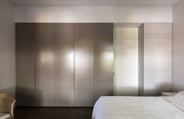 A minimalist bedroom with a large wall of full-height taupe built-in wardrobes, a recessed open wardrobe or dressing area, dark timber flooring, and a partially visible bed with white textured bedding. A woven armchair sits at the left edge. The room has a restrained, contemporary fit-out with clean lines and neutral tones.
