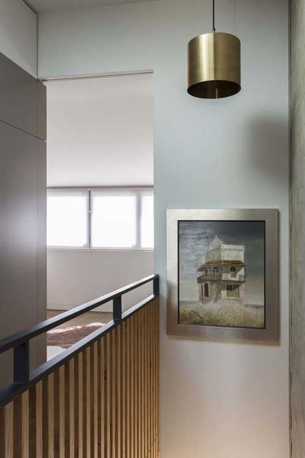 A contemporary interior landing or hallway with a high pale wall, a framed architectural artwork, and a suspended cylindrical brass pendant light. A black-framed timber slat balustrade overlooks a lower level, while soft daylight enters through roller-shaded windows in the background. The palette is restrained, combining pale grey walls, warm timber, black metal, and brass.