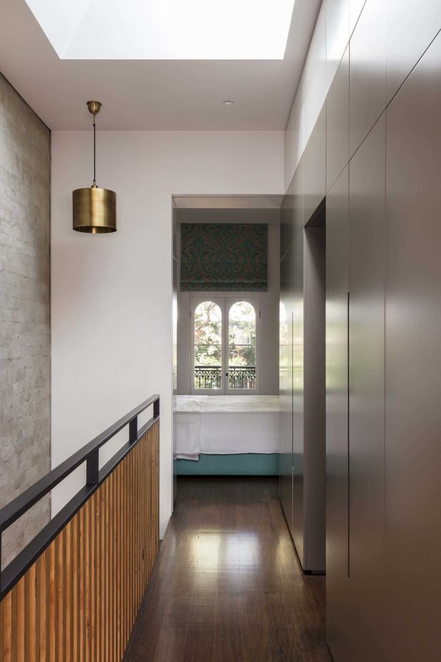 A narrow, refined upper-level hallway or landing with dark timber flooring, a textured exposed brick wall, full-height muted grey built-in cabinetry, and a timber slatted balustrade with black metal framing. A brass pendant light hangs beneath a large skylight. At the end of the corridor, an adjoining bedroom is visible through an opening, featuring a bed, patterned window covering, arched windows, and a balcony beyond.