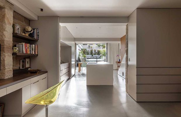 Contemporary open-plan interior viewed from a hallway or entry zone, with polished concrete flooring, full-height grey cabinetry, a compact built-in display and storage unit, and a sculptural yellow chair. The space opens toward a bright kitchen and dining area with an island, while exposed rough stonework adds texture to the otherwise restrained, minimalist fit-out.