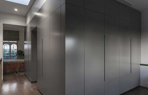 A refined interior hallway or dressing area dominated by a large wall of dark grey, floor-to-ceiling built-in cabinetry with slim vertical handles. Warm timber flooring, white walls and ceiling, recessed lighting, and a skylight create a clean, contemporary look. A glimpse of a richly patterned bedroom or sitting area is visible through the opening at the rear.