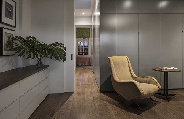Refined contemporary interior hallway or entry zone with dark timber flooring, smooth grey built-in cabinetry, a minimalist white drawer unit, framed artwork, and a sculptural upholstered lounge chair beside a small round side table. A decorative stained-glass or patterned glazed doorway is visible at the end of the corridor, adding a subtle traditional character to the otherwise modern space.