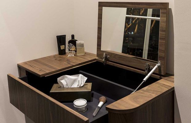 Close-up of a compact, custom dark wood dressing table or vanity with a hinged central mirror and integrated storage compartments. The vanity has black interior surfaces, curved drawer fronts, chrome hardware, and toiletries arranged on the countertop. A window is reflected in the mirror.