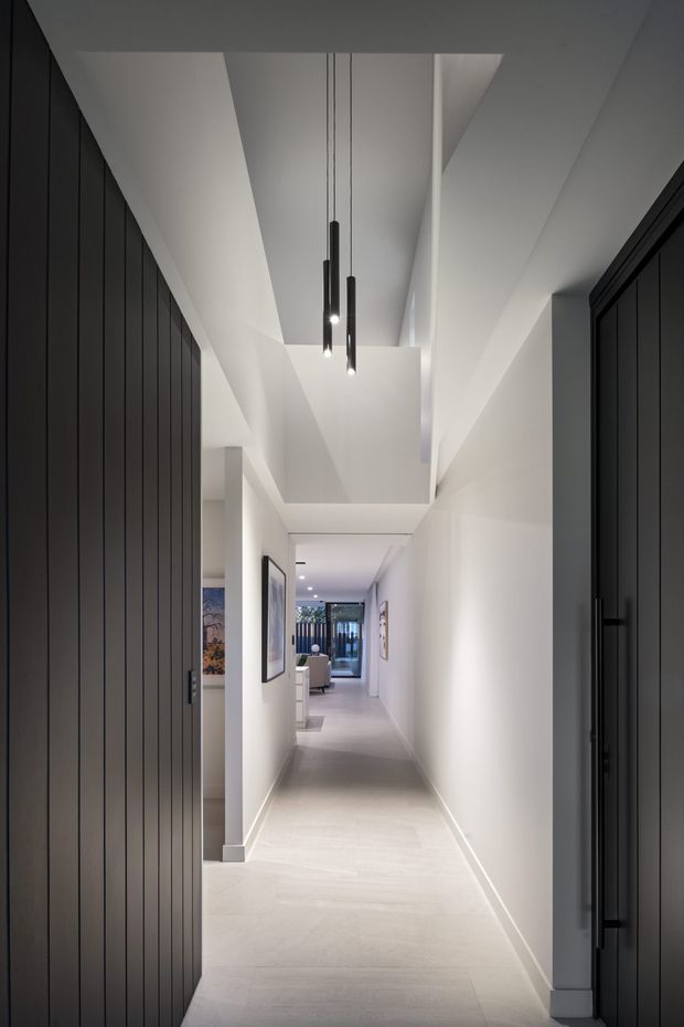 A long, narrow contemporary hallway with white walls, pale flooring, dark full-height timber-panelled doors and cabinetry, and a dramatic stepped ceiling with three slim black pendant lights. Framed artwork and a glimpse of a living area appear at the far end.