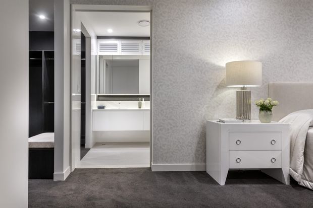A refined bedroom interior viewed toward an open ensuite bathroom. The bedroom features soft grey patterned wallpaper, plush dark grey carpet, a white bedside table with a lamp and floral arrangement, and part of a neatly made bed. The ensuite has white cabinetry, a broad vanity mirror, a white basin, pale flooring, black accent wall panels, and a high louvred window. The overall palette is neutral, bright, and understated.