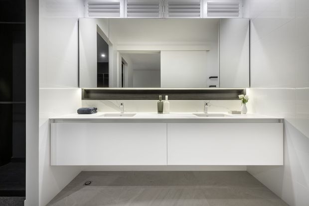 A bright, contemporary bathroom featuring a wide double vanity with white integrated basins, streamlined white cabinetry, a large wall-to-wall mirror, and white tiled walls. Grey stone-look flooring, minimalist fixtures, and high-level louvred ventilation provide a clean, functional finish.