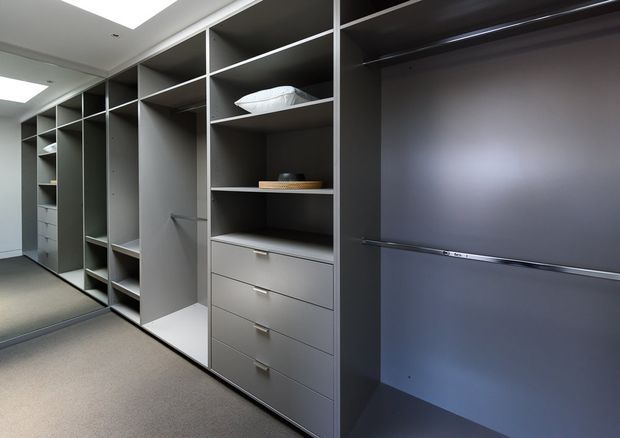 A modern walk-in wardrobe or dressing room with extensive floor-to-ceiling built-in grey cabinetry, open shelving, drawers, hanging compartments, and a large dark wardrobe section. The room has a neutral carpeted floor, white ceiling, recessed lighting, and a clean, minimalist fitted-storage design.