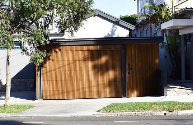 Street-facing view of a contemporary residential garage or carport enclosure with a broad timber slatted garage door and matching pedestrian door, framed in dark metal. The structure has a simple gable roof, white rendered walls, and is integrated beside a multi-storey dwelling with balconies. A driveway, footpath, lawn, and mature planting are visible in front.