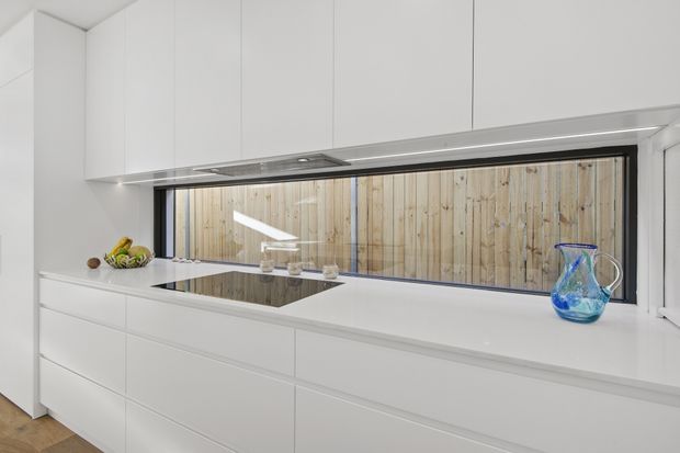 Bright minimalist white kitchen with extensive handleless cabinetry, a long white benchtop, integrated black induction cooktop, and a slim black-framed horizontal window. A timber privacy fence is visible outside the window, while a blue glass jug and small fruit arrangement provide subtle colour accents.