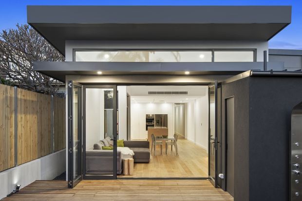 Contemporary two-storey rear facade of a compact residential extension, photographed from a timber deck at dusk. The dark rendered exterior has strong horizontal flat roof forms, deep overhangs, and extensive black-framed glazing with folding or sliding doors. Through the open glazing, a bright open-plan living and dining area with timber flooring, a grey sofa, dining table, and kitchen cabinetry is visible. A timber privacy fence lines the side boundary, and a stainless-steel outdoor kitchen or barbecue is partially visible at right.