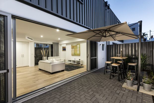 Contemporary courtyard entertaining area viewed from outside, with a large retractable glass opening connecting a paved patio to a bright living room. The exterior features dark vertical cladding, charcoal privacy fencing, light timber flooring indoors, and an outdoor dining setting beneath a large cantilevered umbrella.