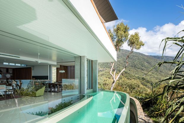 Exterior view of a contemporary elevated home with a covered open-plan living and dining area beside a curved turquoise swimming pool. The house features broad roof overhangs, pale concrete structural columns, extensive glazing, timber screening, and a light soffit. Dense native vegetation and a steep forested mountain backdrop reinforce the hillside setting.