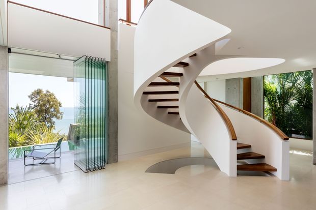 A bright, double-height contemporary interior dominated by a sculptural curved white staircase with dark timber treads and a timber handrail. The space has pale glossy flooring, white walls, tall glazing with vertical glass-panel screening, and views to lush outdoor planting and a terrace. A partial upper-level void and streamlined curved architectural forms create a dramatic, airy entrance or living space.