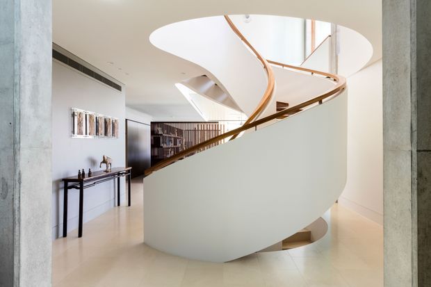A bright, refined interior dominated by a sculptural curved staircase with a smooth white balustrade and warm timber handrail. The stair rises around a large oval opening beneath a skylit or double-height void. Pale flooring and walls are framed by exposed concrete columns, while a slim dark console and artwork sit in the background.