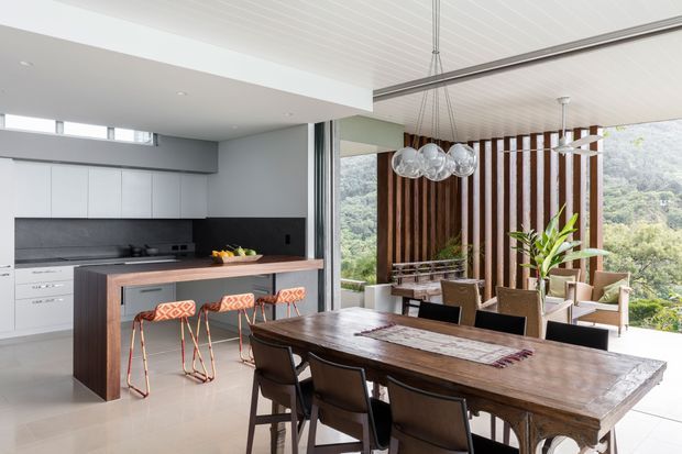 Open-plan contemporary kitchen and dining area with a large rustic timber dining table in the foreground. The kitchen has sleek white cabinetry, a dark stone-look splashback and island, while the dining zone features a vertical timber screening wall, sculptural glass pendant lights, and expansive glazing overlooking a green hillside.