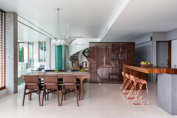 Open-plan contemporary residential interior combining a dining area, kitchen and living zone. A long timber dining table with upholstered chairs sits beneath sculptural pendant lights, while a timber kitchen island with bar stools occupies the right side. Vertical timber screening partially separates the kitchen from a curving staircase and living area. The space features extensive glazing, concrete columns, a white linear-panel ceiling, polished pale flooring and warm timber accents.
