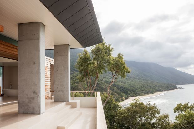 A contemporary concrete-clad terrace or balcony projects from a modern home on a steep coastal site. The view looks over a forested mountain slope, sandy shoreline, and calm bay beneath an overcast sky. Broad concrete columns, pale tiled or stone steps, timber wall cladding, a dark metal roof edge, and a partially visible bathroom or interior space create a refined indoor-outdoor architectural composition.