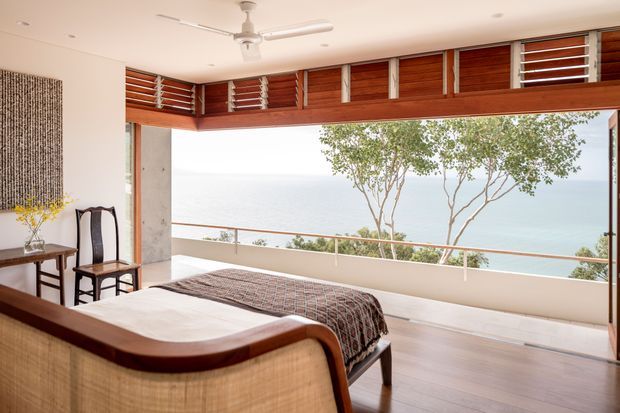 A bright coastal bedroom opening onto a broad ocean-facing balcony or terrace. The room features a low wooden bed with a patterned throw, a curved woven-back bench or bed-end seat, two dark wooden traditional chairs, a small side table, and a large textured wall hanging. Full-width timber-framed glazing incorporates horizontal louvres above the opening, while a ceiling fan and pale timber flooring reinforce the airy tropical setting. Ocean and trees are visible beyond the balcony.