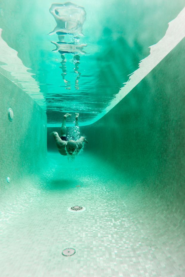 Underwater view inside a tiled swimming pool, looking along the pool floor toward a submerged swimmer. The walls and floor are covered in small pale aqua mosaic tiles, with pool drains and fittings visible. The water creates strong reflections and rippling patterns on the surface, producing a monochromatic turquoise atmosphere.