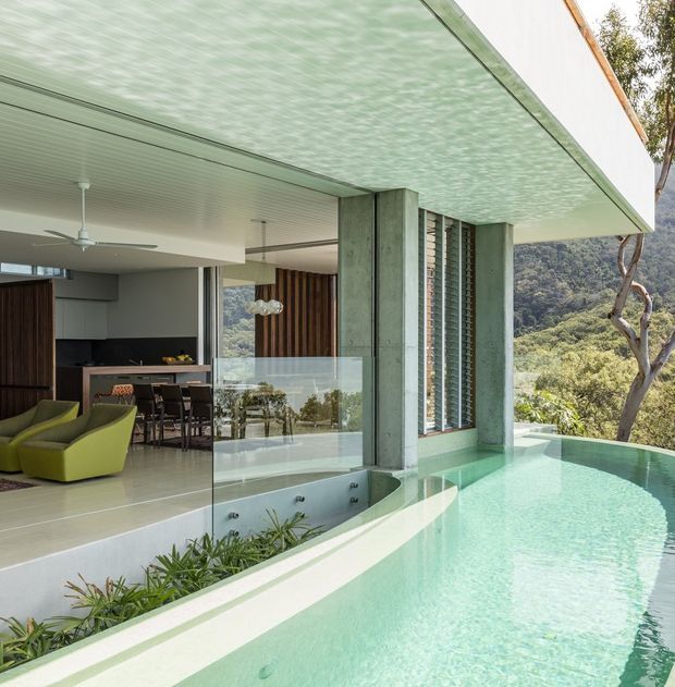 A contemporary open-plan living and kitchen area opens directly onto a narrow infinity-edge swimming pool. The image highlights a broad cantilevered flat roof, exposed concrete columns, floor-to-ceiling glazing, vertical timber screening, and a transparent glass balustrade. Lime-green lounge chairs and a dining setting sit beside the kitchen, while a wooded hillside is visible beyond the glazing.