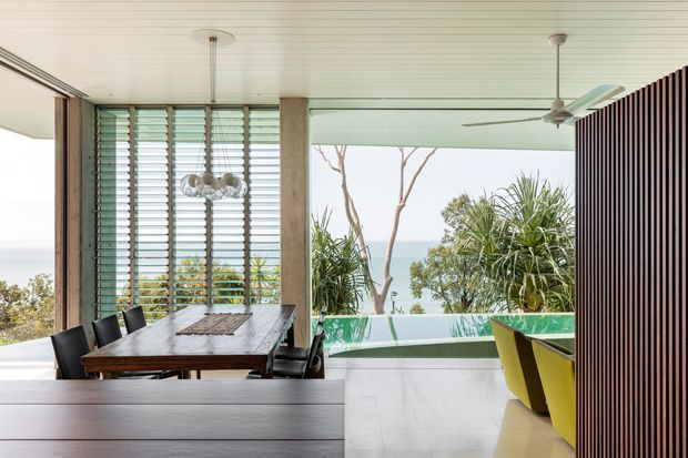 A bright, open-plan coastal dining and living area with expansive glazing and ocean views. A dark timber dining table with black chairs sits beside louvred windows, while a sculptural pendant hangs above. The space features pale flooring, a white slatted ceiling, a ceiling fan, a green-tinted glass balustrade, and a bold vertical timber screen separating the adjacent seating area with yellow chairs. Tropical planting is visible beyond the glazing.