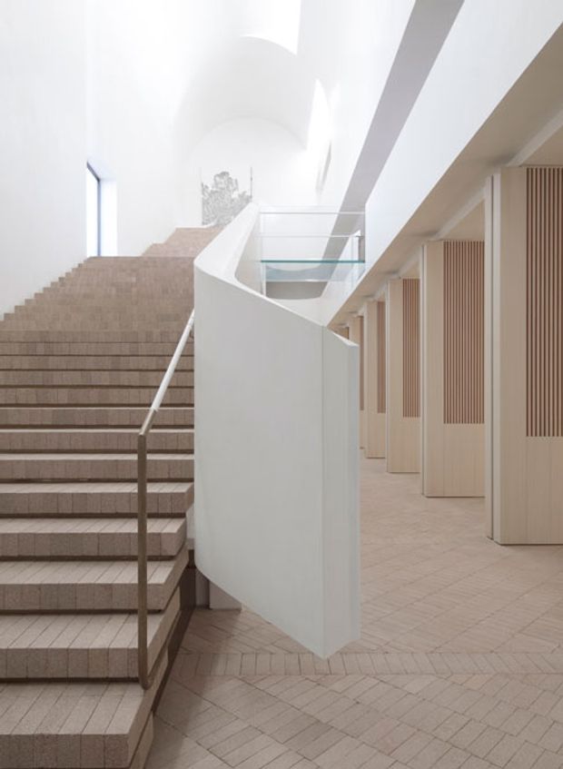 A bright, refined interior stairway and landing with broad pale stone or porcelain-tiled steps, a slim metal handrail, and a sculptural white curved stair wall. The adjoining corridor features pale vertical timber slat panels and built-in doors, while a glazed balustrade and high white ceiling create an airy, gallery-like atmosphere. The floor uses a repeating herringbone or chevron tile layout.