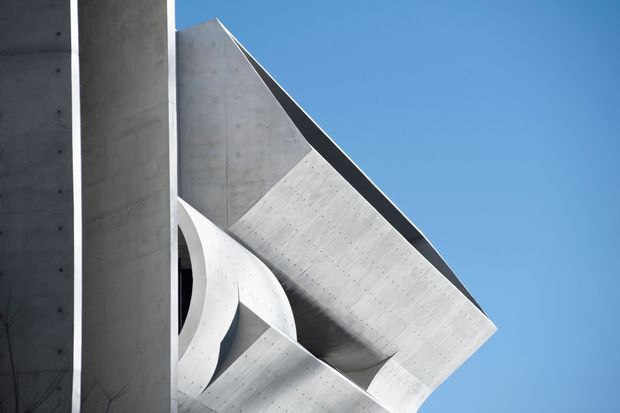 Abstract close-up of a sculptural modern concrete building facade, showing sharply folded geometric planes, a sweeping curved concrete element, and a tall vertical wall against a clear blue sky. The image emphasizes raw materiality, dramatic angles, and monumental architectural form.