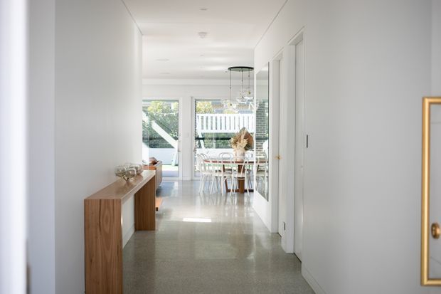Bright, narrow residential hallway leading toward a dining area and glazed exterior doors. The space has white walls and doors, recessed ceiling lights, polished light-grey flooring, a timber console along the left wall, and a large framed mirror on the right. A dining table with white chairs is visible at the end of the corridor.