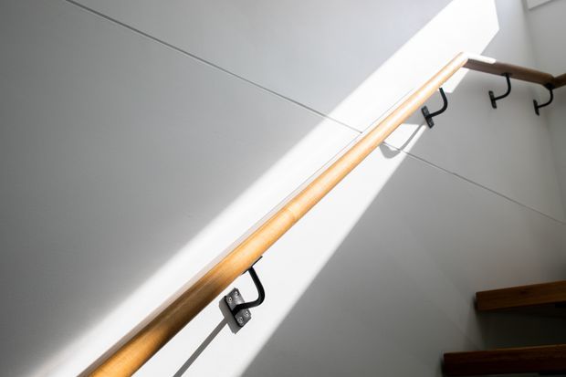 Close-up architectural view of a timber handrail mounted along a white stairwell wall, with black metal brackets and coat-style hooks. Strong sunlight creates a diagonal beam across the smooth wall, while the edge of dark timber stair treads is visible at lower right.