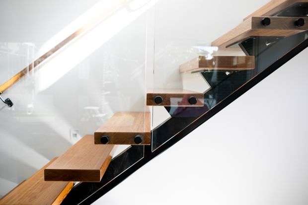 Close-up view of a modern floating staircase with thick timber treads cantilevered from a dark structural stringer. A clear glass balustrade with black circular fixings runs alongside the stair, set against white walls and bright natural light.