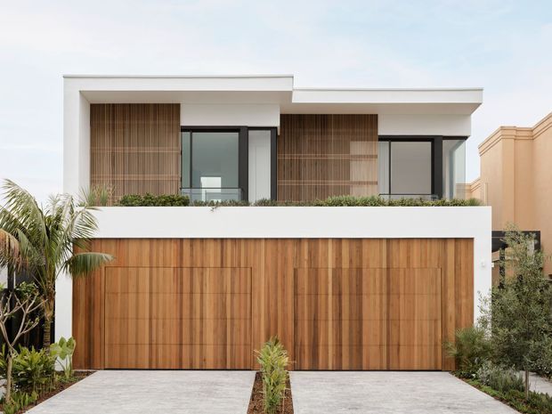 Contemporary two-storey residential facade with clean white rendered planes, a flat roofline, extensive dark-framed glazing, and warm vertical timber cladding across the upper-level screens and large garage doors. A low planted balcony edge and tropical landscaping soften the geometric composition, while pale concrete driveways lead to the two-car garage.