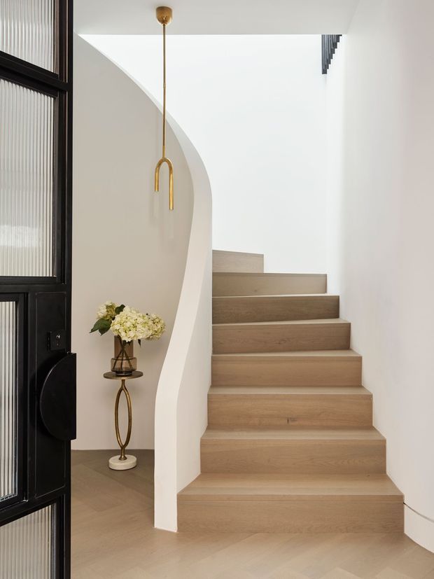 A refined interior stairway with broad pale timber stairs rising beside a sculptural curved plaster wall. A black-framed glazed entry door is visible at left, while a slender brass pendant light and a small floral arrangement add understated decorative detail. The space is bright, minimal, and softly neutral.