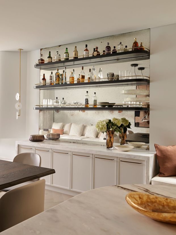 A refined dining and entertaining area with a large mirrored built-in bar cabinet, three open glassware and liquor shelves, white paneled cabinetry, and a pale stone countertop. A dark timber dining table and upholstered chairs occupy the foreground, while a built-in upholstered bench with cushions and floral arrangements sits beneath the bar. The space has soft neutral finishes, warm lighting, and elegant contemporary detailing.