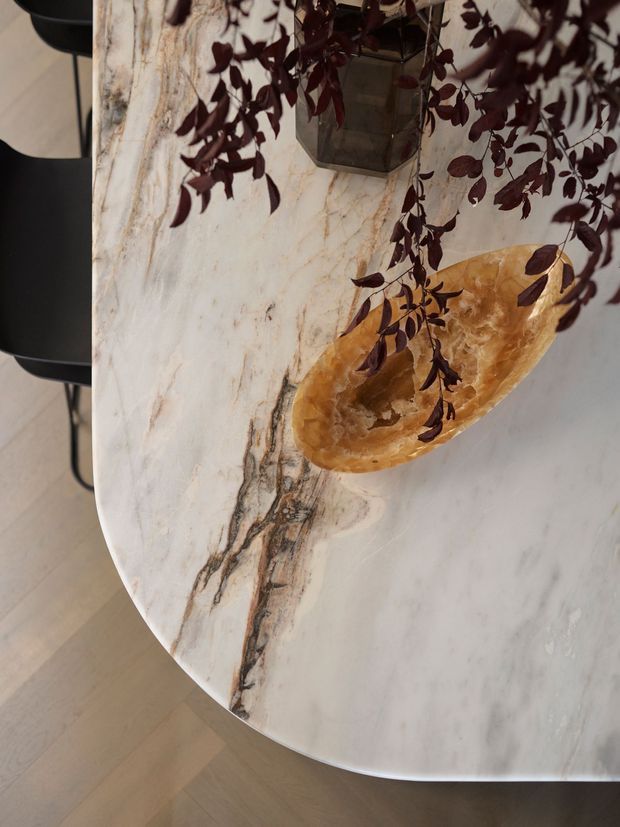 Close-up of a rounded-edge dining table with a dramatic white marble surface featuring grey and warm brown veining. A golden-brown organic stone or geode bowl sits on the tabletop alongside a dark hanging planter, with black dining chairs partially visible around the table. Light timber flooring is visible below.