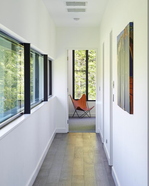 Bright, narrow residential hallway with white walls and ceiling, a long strip of black-framed glazing on the left, pale grey rectangular floor tiles, and a colourful wall artwork on the right. The corridor leads to a small sunlit sitting area with a modern butterfly chair and large window overlooking trees. Recessed linear ceiling vents and simple white door trims contribute to a clean contemporary feel.