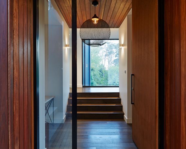 A refined residential entry hall viewed through an open timber door, featuring a dark timber floor and stairway, warm vertical timber wall cladding, a timber-lined ceiling, white walls, black-framed glazing, and sculptural pendant lighting. The stair leads toward a large window overlooking leafy vegetation, creating a warm contemporary interior with strong natural-material detailing.