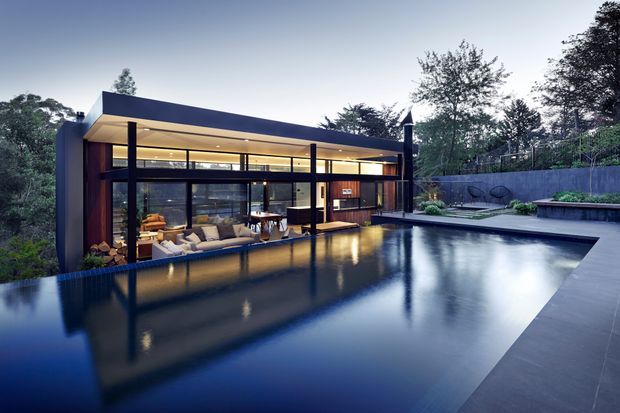 Contemporary single-storey house with a low, flat roof and extensive floor-to-ceiling glazing opening onto a large reflective swimming pool. The dark facade and slender structural posts contrast with warm interior lighting, while a paved pool terrace, raised garden beds, boundary wall and mature trees frame the setting.