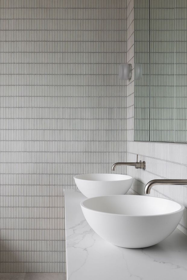 A refined contemporary bathroom featuring two white vessel basins on a pale marble-look vanity, paired with brushed metal wall-mounted taps. The walls are fully clad in fine vertically ribbed light-grey tiles, with a large mirror above the basins. The design is monochromatic, clean-lined, and minimalist.