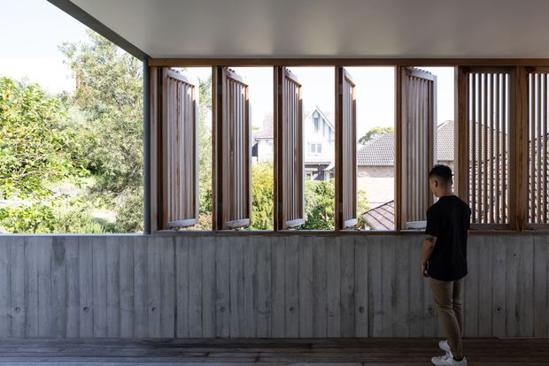 A covered architectural space with a long row of timber-framed louvred openings and vertical timber screens above a textured grey concrete half-wall. The open panels overlook leafy suburban surroundings and neighbouring rooftops, while a person stands near the right side. The space is bright and airy, with strong linear detailing and a warm timber-and-concrete material palette.