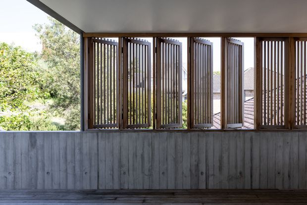 A covered outdoor architectural space featuring a row of vertical timber louvres set within dark timber frames above a weathered concrete or grey cement-panel half wall. The louvres provide filtered views to leafy vegetation and neighbouring rooftops, while a timber deck or floor and flat soffit complete the restrained, natural-toned design.