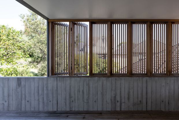 A covered outdoor veranda or terrace with a low vertically board-formed concrete wall, dark timber decking, and a series of timber-framed vertical slatted screens. The screens filter views of lush vegetation and a neighbouring roof beyond, creating a shaded, private architectural space. The composition is minimalist and strongly defined by the repetitive timber louvers and concrete surfaces.