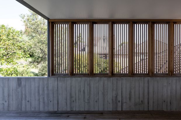 A covered outdoor architectural space with a dark timber slatted screen above a weathered concrete or grey timber-clad low wall. The screened openings look onto leafy vegetation and neighbouring roof forms, while a smooth light ceiling and dark deck frame the composition. The image emphasizes privacy, filtered light, and rhythmic vertical detailing.